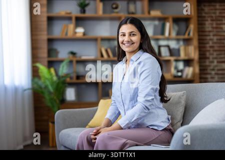 Young indian woman feeling confident and cheerful, sitting on a comfortable sofa in a stylish living room or home office background, making eye contact and presenting a friendly expression Stock Photo
