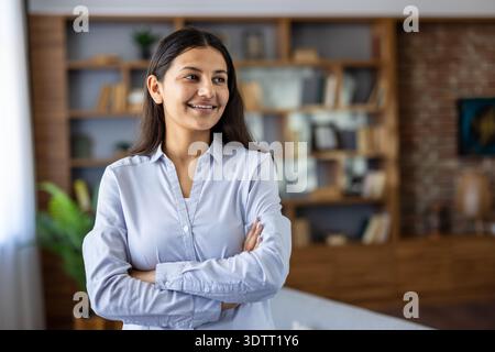 Young confident indian woman standing with arms crossed, smiling and looking away in a modern home office or library with bookshelves and exposed brick, radiating leadership and optimism Stock Photo