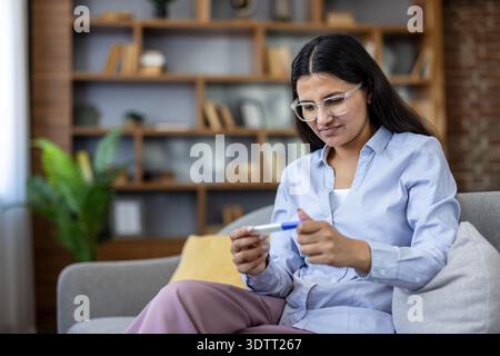 Young woman sitting on a couch at home, looking at a pregnancy test with anticipation, experiencing emotions of hope and uncertainty about family planning Stock Photo