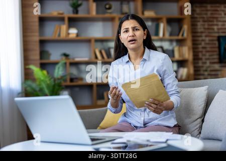 Young woman sitting on a sofa at home, opening an urgent envelope with official documents and a laptop nearby, feeling stressed about unexpected financial problems Stock Photo