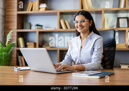 Young indian woman wearing eyeglasses smiling at the camera, working on a laptop at a wooden desk in a modern office with bookshelves in the background Stock Photo