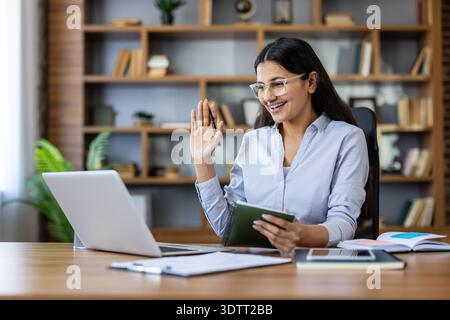 Young smiling woman wearing glasses engaging in a video conference with a laptop, holding a notebook and pen, working remotely from a home office or modern workspace Stock Photo