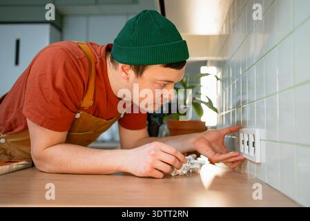 Handyman installing new multiple electrical sockets into a tiled kitchen wall, wearing work overalls and a green beanie, ensuring a precise and secure fit for home electrical renovation Stock Photo