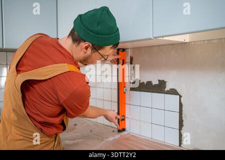 Skilled handyman carefully applying ceramic tiles to a kitchen wall, covering the surface with adhesive and checking alignment using a spirit level for a precise installation Stock Photo
