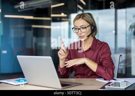 Young businesswoman wearing a headset and glasses, actively participating in a virtual conference or customer support call from her office desk using Stock Photo