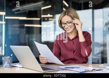 Young professional businesswoman smiling, correcting glasses, and analyzing documents and financial reports while sitting at her office desk with a la Stock Photo