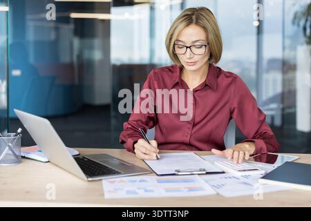 Professional woman wearing glasses balancing finances, managing documents, and calculating expenses on a desktop with a laptop in a modern office envi Stock Photo