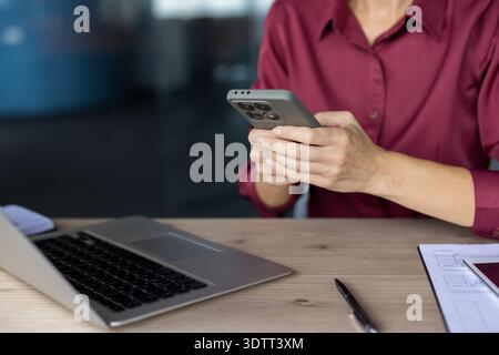 Businesswoman's hands holding and interacting with a smartphone, with a laptop, pen, and documents visible on a wooden desk in a modern office environ Stock Photo