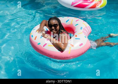 Woman floating on donut inflatable with sprinkles in outdoor pool in red swimsuit and sunglasses Stock Photo