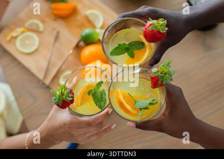 Three citrus cocktails are clinking on wooden table, with mint, orange, strawberry garnishes Stock Photo