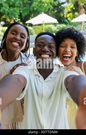 African friends posing and taking smartphone selfie at sunny poolside with white umbrellas Stock Photo