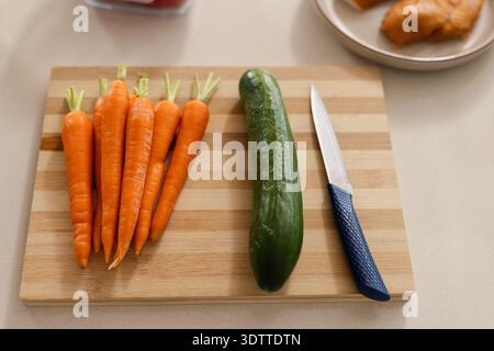 Wooden cutting board is holding seven carrots, cucumber and knife on light counter, pastry blurred Stock Photo