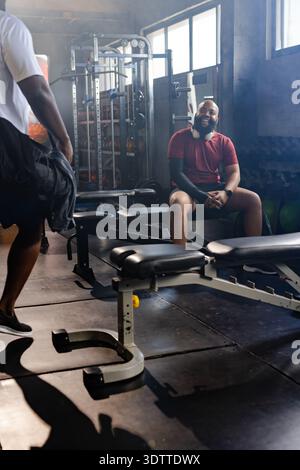 African male friends sitting on bench in gym weight area, smiling with headphones, copy space Stock Photo