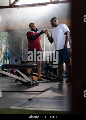 Two African American men fist-bumping while training in gym near weight bench, wearing shorts Stock Photo