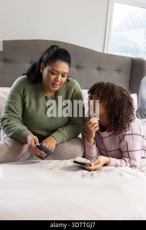 African American female mother and teenage daughter sitting on bed in bedroom holding smartphones Stock Photo