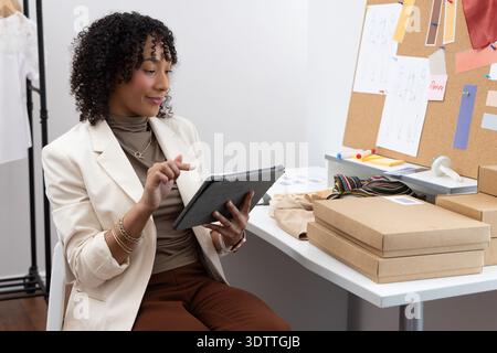 African American woman wearing cream blazer sitting at desk using tablet, packing boxes, copy space Stock Photo