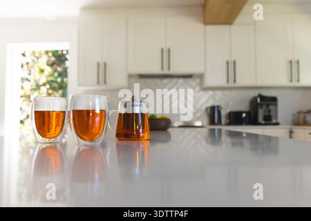 Two double-walled glass cups and glass teapot are reflecting amber tea on glossy white island Stock Photo