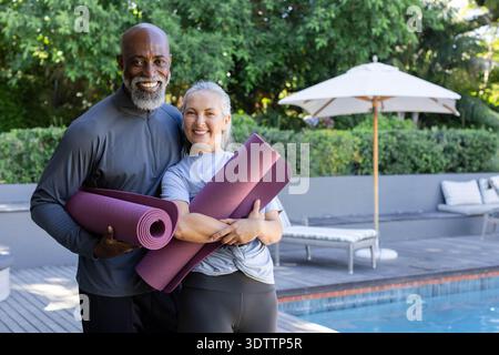 Diverse senior couple standing together on wooden pool deck holding two purple yoga mats, smiling Stock Photo