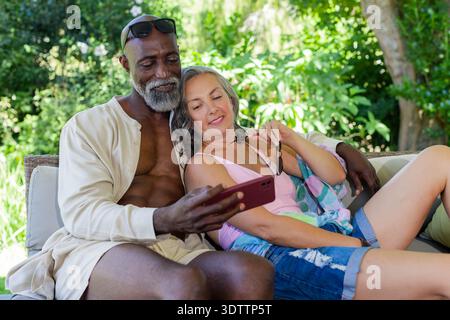 Diverse couple reclining on cushioned outdoor sofa in garden, checking red smartphone together Stock Photo