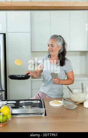 Senior woman flipping pancake in black frying pan on wooden countertop with gray spatula nearby Stock Photo