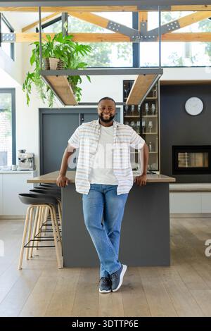 African man standing and leaning on central kitchen island, smiling near coffee machine and plants Stock Photo