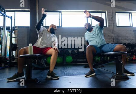 Two African American men highfiving sitting on benches in gym with dumbbell rack and water bottle Stock Photo