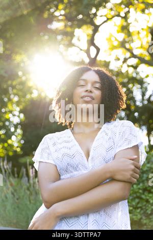 African American woman standing arms crossed in sunlit garden with white eyelet dress, rim light Stock Photo