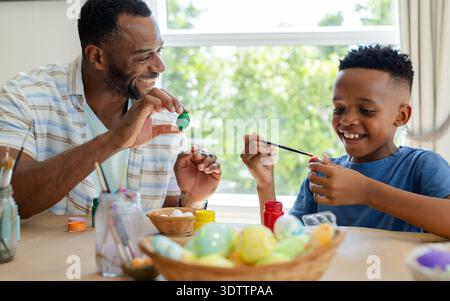 African American father and child painting eggs with paint jars at wooden table by window Stock Photo