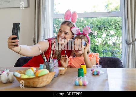 Mother in red top, daughter wearing pink bunny headbands painting eggs at home taking phone selfie Stock Photo