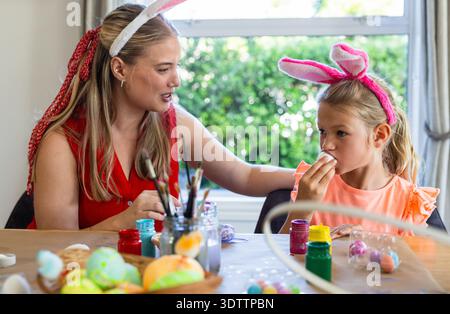 Woman and child wearing bunny ears decorating eggs at home table with paints, brushes, painted eggs Stock Photo