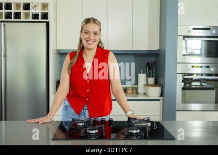 Adult woman leaning on stone island in modern kitchen, smiling in red top, showing cooktop Stock Photo
