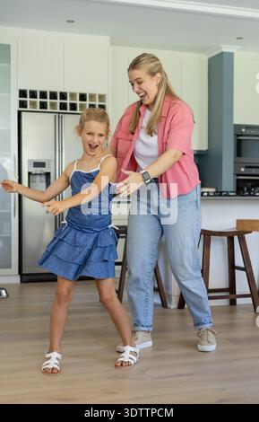 Mother and daughter dancing in kitchen near fridge, mother wearing smartwatch, child in blue dress Stock Photo