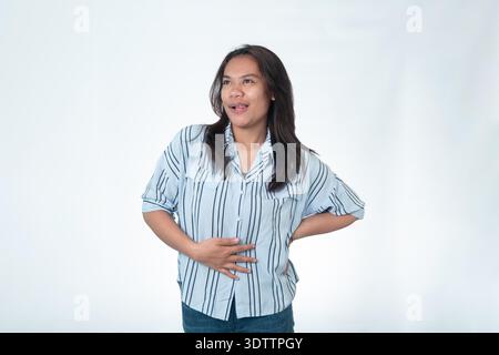 Indonesian woman in a blue striped shirt looking aside with a playful expression, touching her belly with one hand and licking her lips as if satisfie Stock Photo