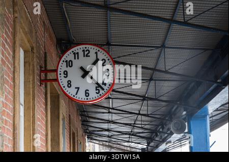 Station clock under metal canopy displaying redrimmed face, mounted on brick station wall with exposed steel beams and corrugated roof, empty platform Stock Photo