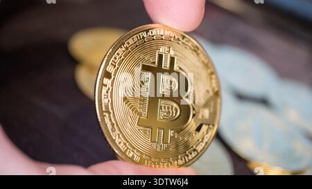 Close-up of a hand holding a shiny gold bitcoin coin with blurred cryptocurrency coins in the background, symbolizing digital finance and investment. Stock Photo