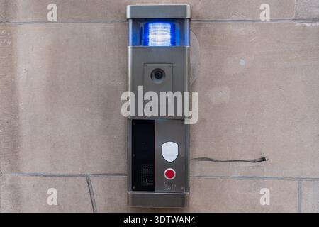 Modern security intercom panel mounted on a stone wall. Stainless steel emergency call box with surveillance camera, speaker, red call button and blue Stock Photo