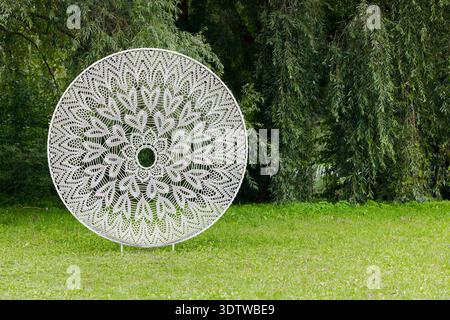 A large, white, circular art installation featuring delicate lace-like patterns stands outdoors amidst vibrant green foliage and grass Stock Photo