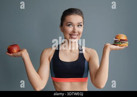 A woman stands with an apple in her left hand and a burger in her right, smiling confidently. She wears a sporty outfit and is set against a plain bac Stock Photo