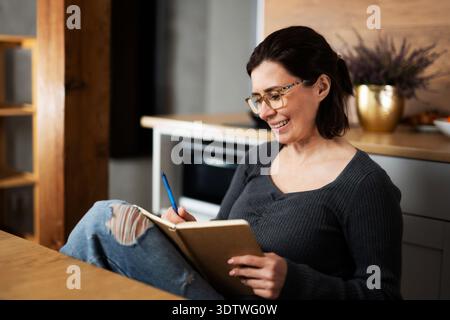 Happy Young Woman Writing Notes In A Notepad In The Kitchen Stock Photo