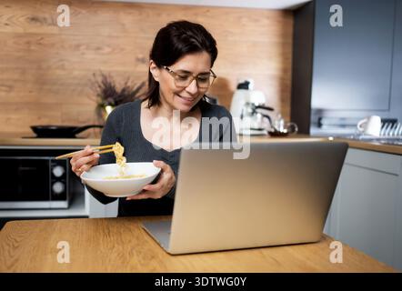 Happy Woman Eating Noodles And Watching A Video On Laptop At The Kitchen Table Stock Photo