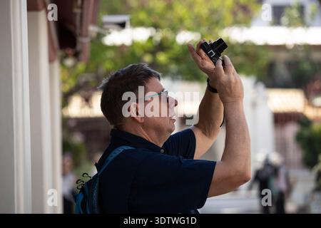BANGKOK, THAILAND-JAN 23, 2026: Senior male tourist taking a photo with a compact camera, face and hand skin texture, side profile Stock Photo