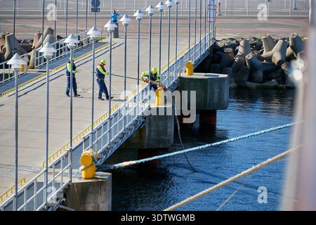 Three dockworkers, dressed in hard hats and high-visibility workwear, are actively engaged in maritime operations on a concrete pier in Tallinn, Eston Stock Photo