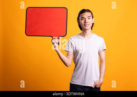Young asian man stands confidently holding blank placard while posing with one hand in pocket. Focused male model holds signboard, presenting clear copy space for business or promotional ads. Stock Photo