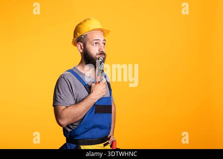 Caucasian male contractor holds pliers thoughtfully under chin, symbolizing planning or repair decision. Bearded renovation worker stands in studio wearing helmet against vibrant yellow background. Stock Photo