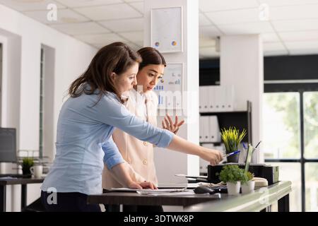Businesswomen stand together at office desk, reviewing project details and discussing strategy. Caucasian and asian coworkers examine information on laptop screen, communicating ideas and next steps. Stock Photo