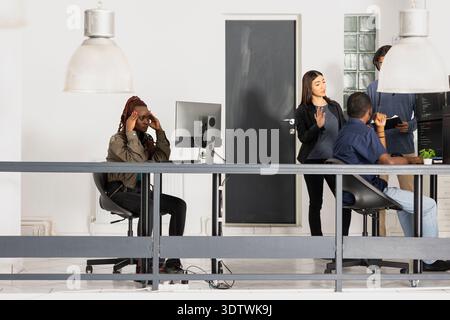 Black woman appears overwhelmed while working in diverse workspace, struggling to complete programming projects. Female developer pauses to ponder debugging coding scripts to achieve a functional app. Stock Photo