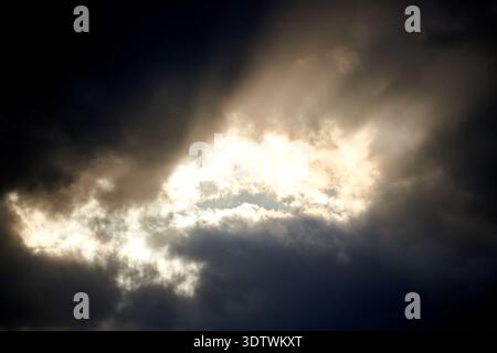 A dramatic sky scene where bright sunlight pierces through a dense formation of dark, ominous clouds. The strong contrast between the brilliant white Stock Photo