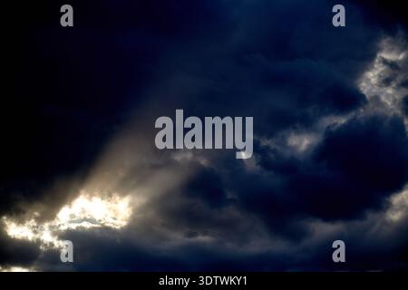 A dramatic and powerful shot of an overcast sky dominated by heavy, dark blue-gray storm clouds. A brilliant ray of light breaks through a gap in the Stock Photo