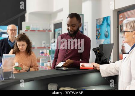 African american man pays for pharmacy items using smartphone, demonstrating cashless payment methods. Senior female pharmacist provides efficient customer service as black customer purchases medicine Stock Photo