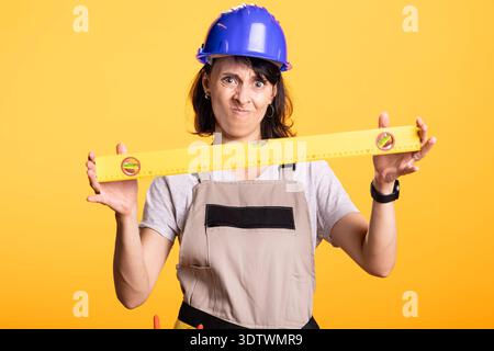 Female builder grips measuring level, questioning accuracy of measurement. Woman contractor angrily holds construction tool, representing frustration in DIY adjustments and onsite evaluation. Stock Photo